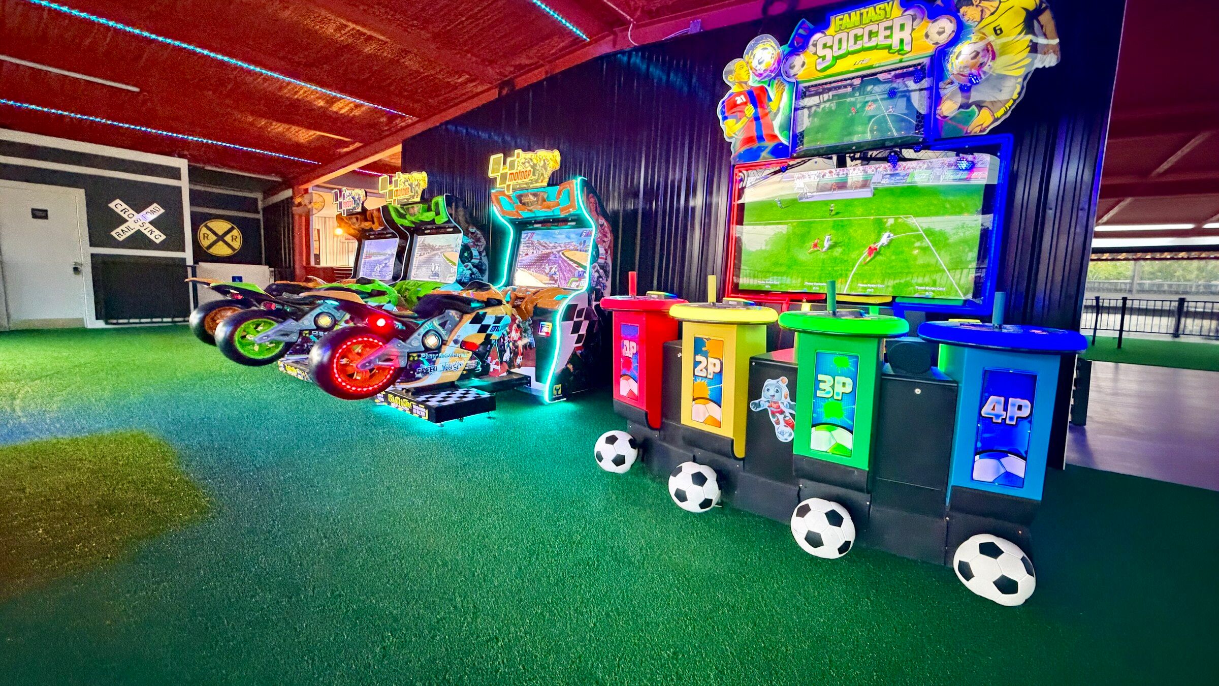 A row of MotoGP riding cabinets beside a four-player Fantasy Soccer console on green turf