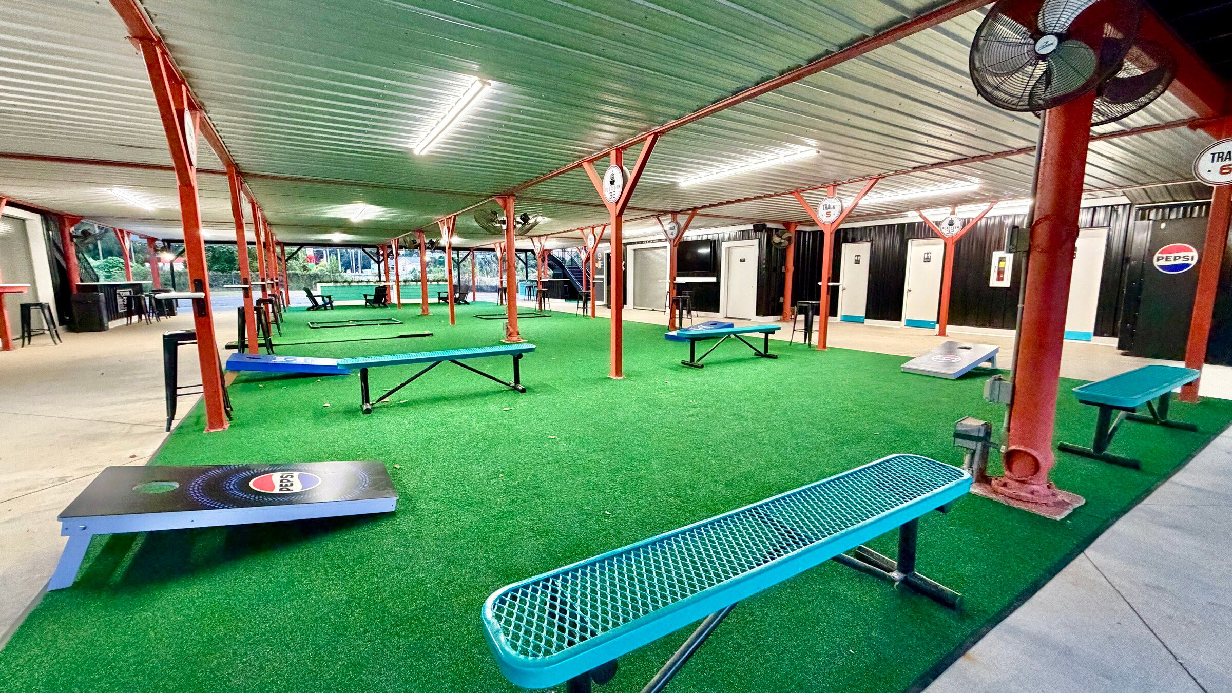 The covered cornhole lawn under Platform 32, Pepsi-branded boards and teal benches on green turf