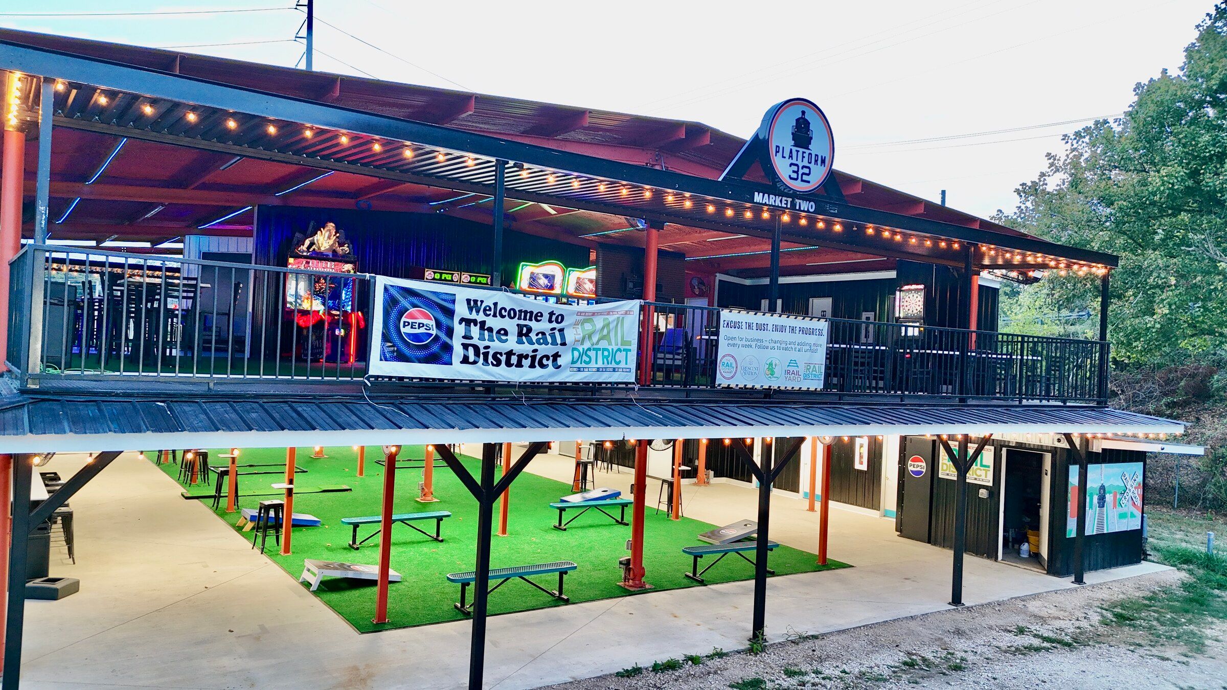 Platform 32 from the air at dusk, the lit arcade on the upper deck and the cornhole lawn on the turf below.